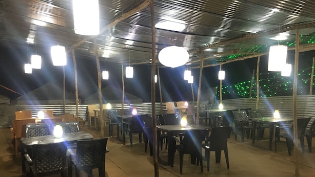An outdoor restaurant with tables and chairs arranged under a covered area with hanging lights. - Kumbh Luxury Tents (Prayagraj)
