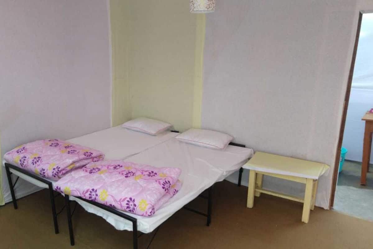 Uma cama de solteiro com colchão branco e duas almofadas, um pequeno banco de madeira e um aplique de parede.