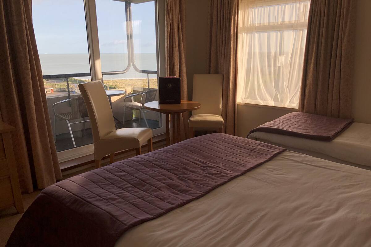 A hotel room with two beds, a laptop on a small table, a chair, and a view of the sea.