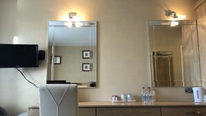 Quarto de hotel com televisão de tela plana na parede, um espelho grande sobre a cômoda e uma cama com cabeceira branca.