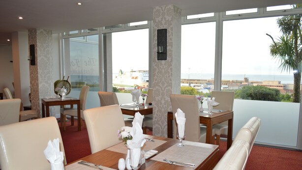 Uma área de refeições com vista para o mar, com uma mesa de madeira posta com guardanapos brancos, talheres e um vaso de flores.