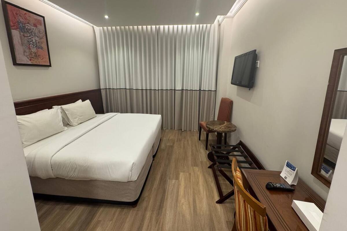 A hotel room with a bed, a chair, a table, a TV, and a window with curtains.