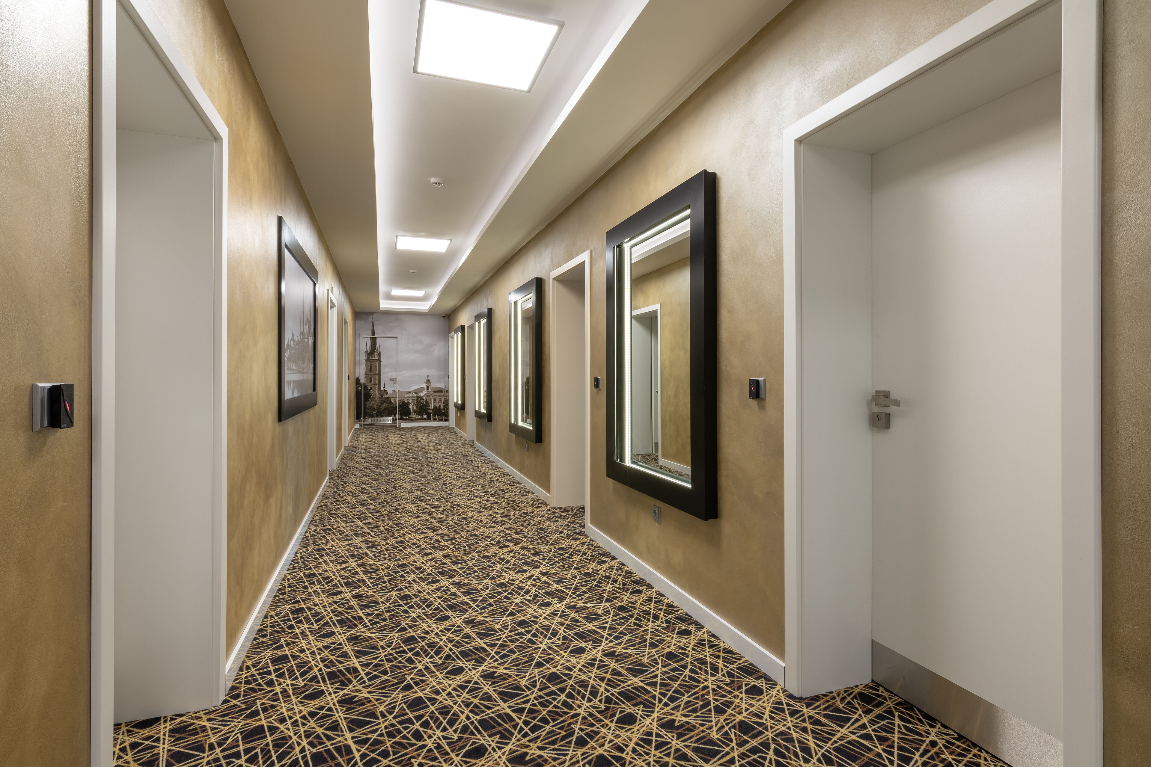 A hotel hallway with patterned carpet, framed mirrors, and doors leading to rooms.