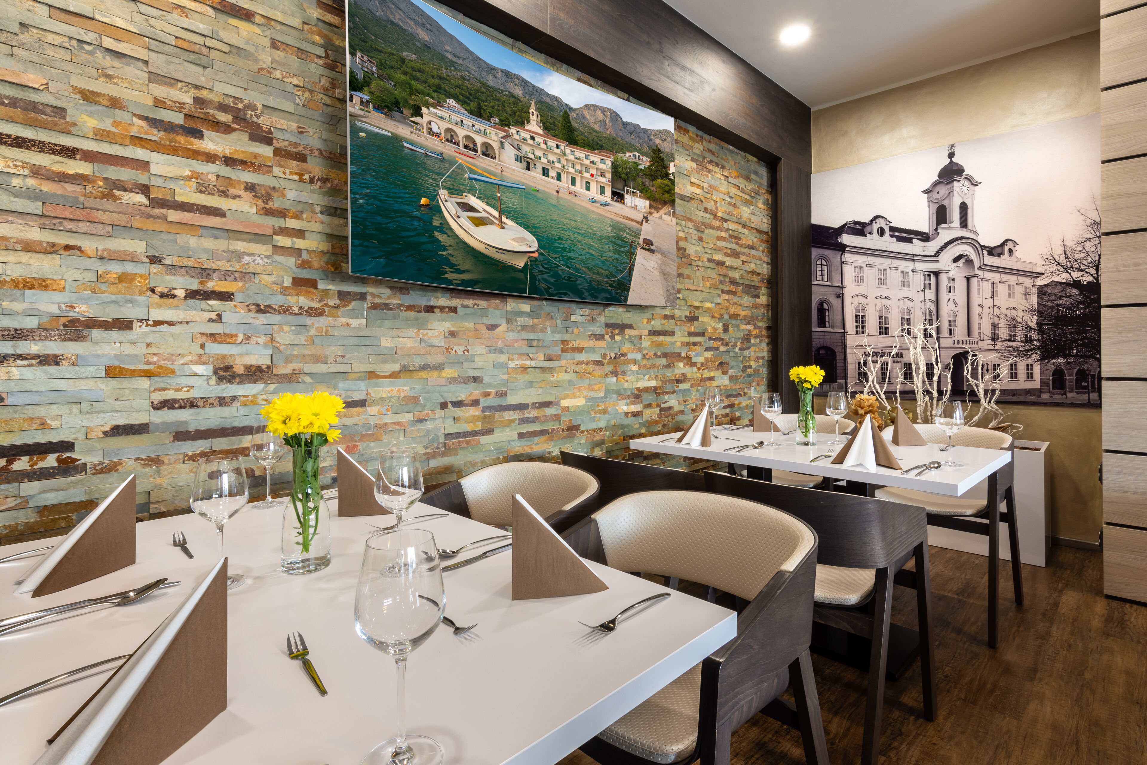 A restaurant with a stone wall and a large painting of a coastal scene. Tables are set with wine glasses and cutlery.