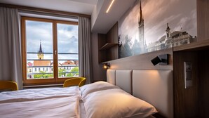 A hotel room with a large bed, a desk, and a view of a cityscape through a large window. - Hotel GRAND (Caslav)