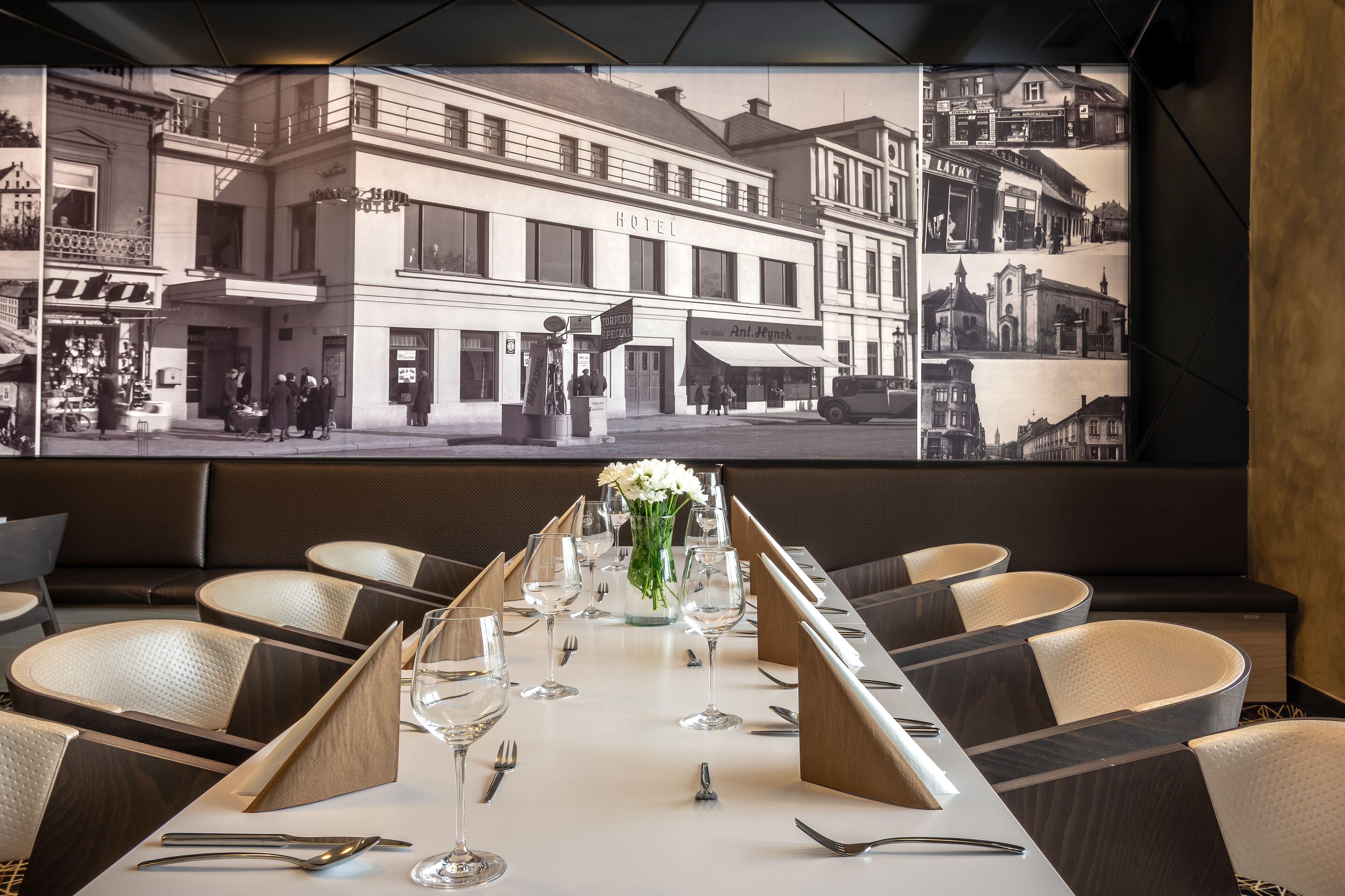 Un restaurante con una larga mesa dispuesta para comer, un amplio ventanal con vistas a un edificio histórico y una exhibición de fotografías blanco y negro en la pared.
