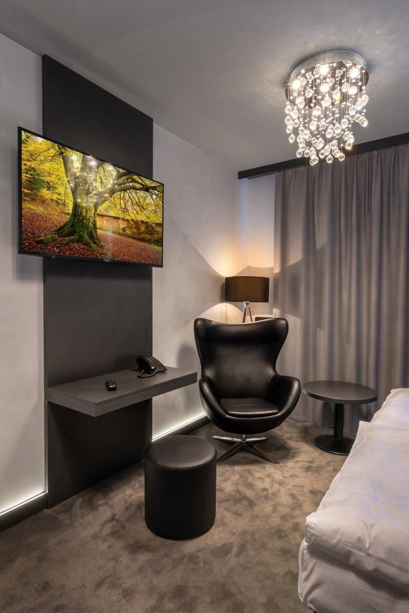 A modern hotel room with a flat-screen TV mounted on the wall, a black leather chair, a small round table, a round ottoman, and a chandelier.