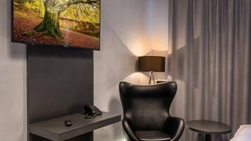 A modern hotel room with a flat-screen TV mounted on the wall, a black leather chair, a small round table, a round ottoman, and a chandelier.