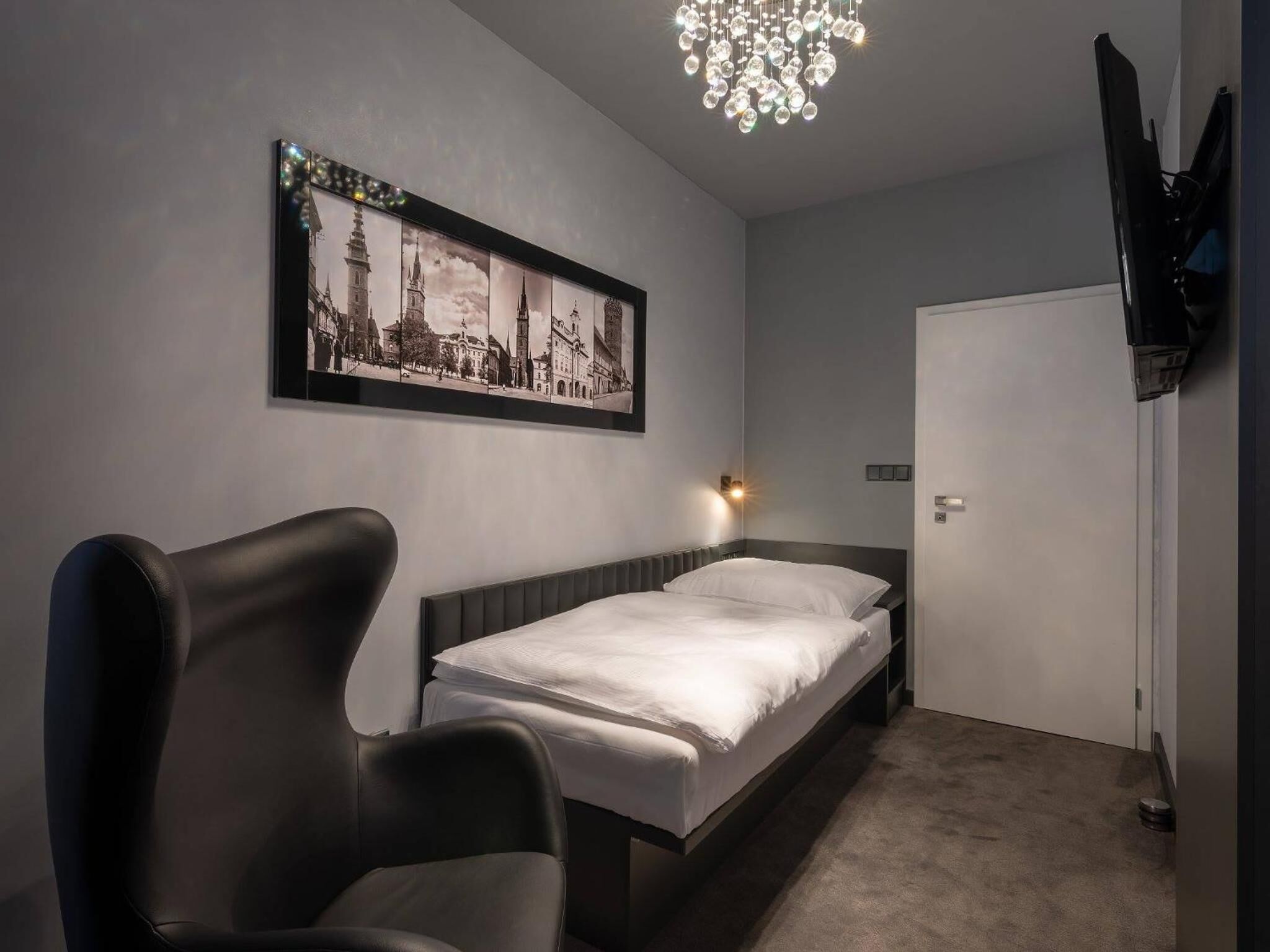 A bedroom with a bed, a chair, a wall-mounted TV, a chandelier, and a framed picture of a cityscape.