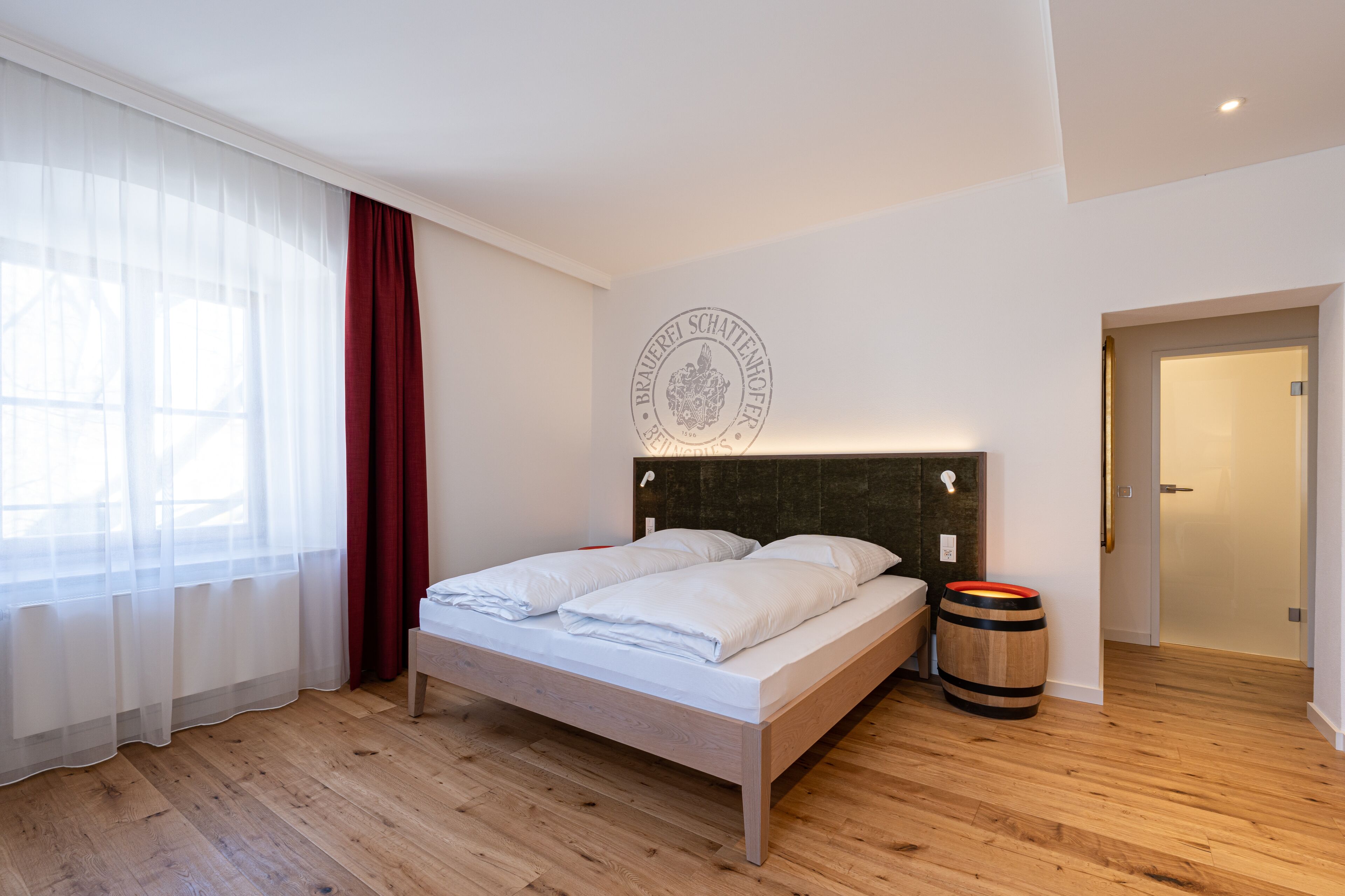 A bedroom with a wooden floor, a bed with white bedding, a wooden headboard, a wall-mounted light, and a barrel.