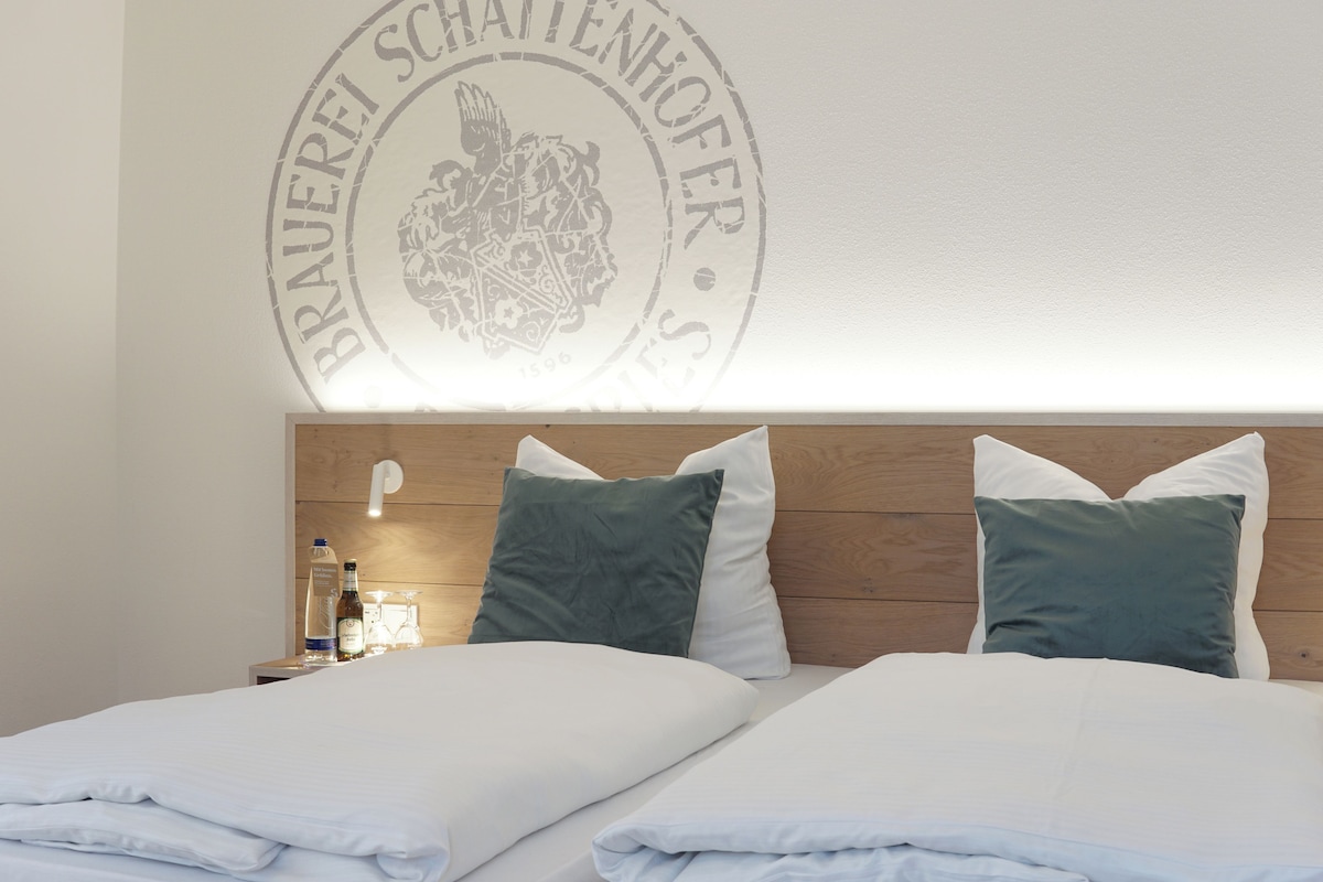 Uma cama de casal bem arrumada com lençóis brancos, duas almofadas verdes e uma cabeceira de madeira. Uma pequena mesa com uma garrafa e um copo é visível à esquerda.