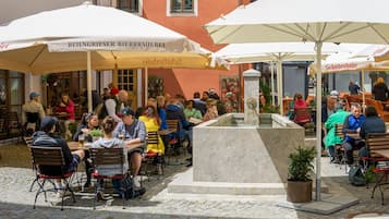 An outdoor café with tables and chairs under umbrellas, a central fountain, and people dining.