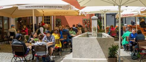An outdoor café with tables and chairs under umbrellas, a central fountain, and people dining.