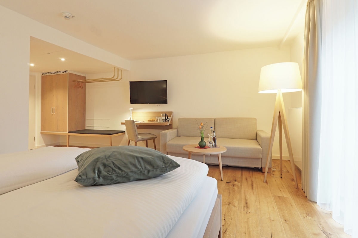 A modern hotel room with a large bed, a sofa, a desk with a chair, a TV, and a lamp.