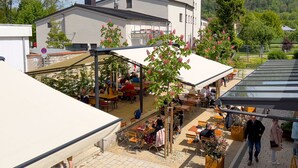 An outdoor seating area with tables and chairs, a building with a sign reading "Schattenhofer," and a view of a forested hill in the background.