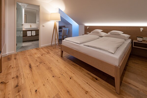 A bedroom with a wooden bed, a bedside table, a nightstand, a lamp, a mirror, and a bathroom with a sink and towel rack. - Braugasthof Schattenhofer (Beilngries)