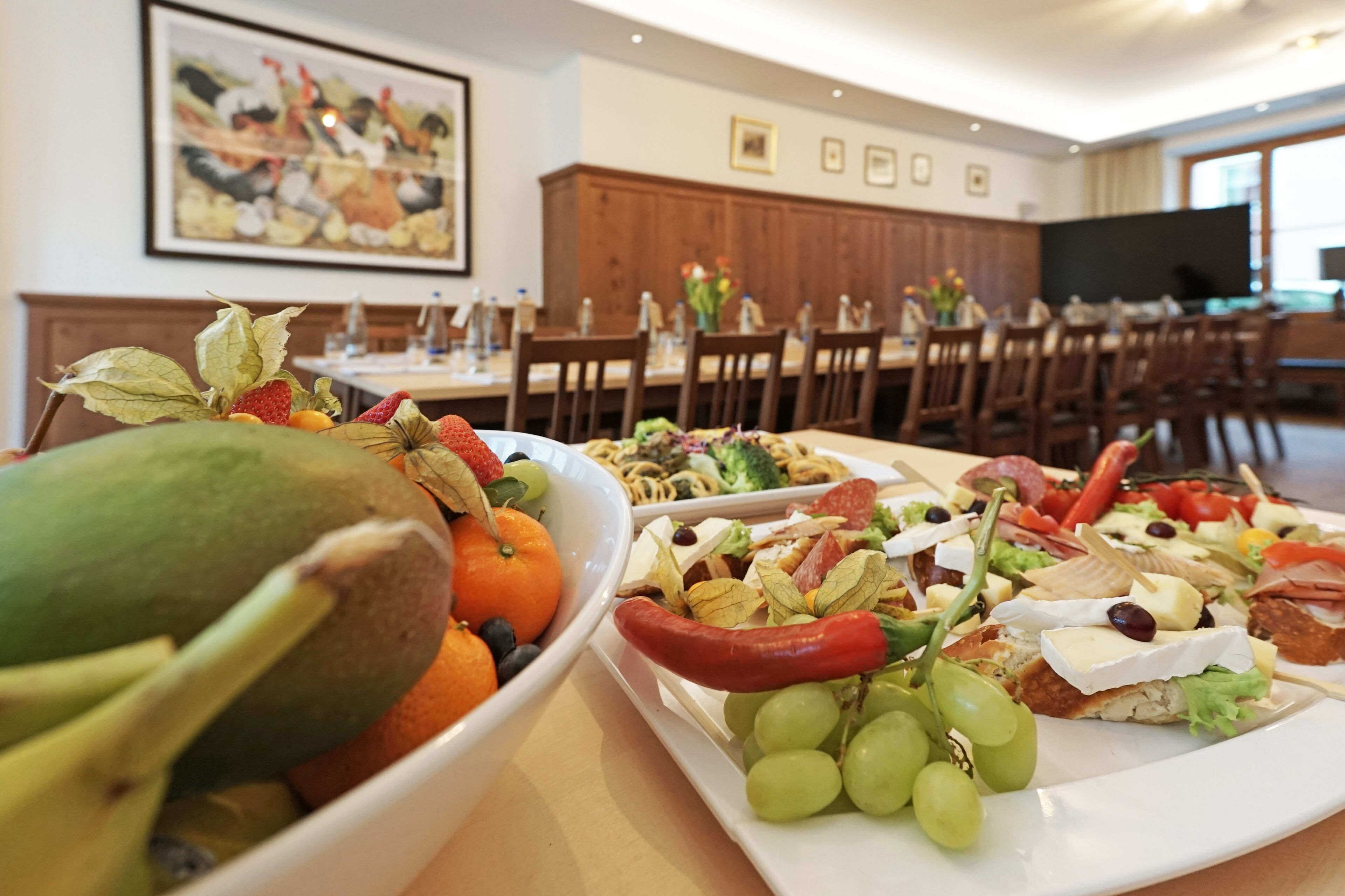 Un espace repas où l’on trouve une variété de plats, notamment des fruits, des légumes et des fromages. On y trouve des chaises et des tables en bois, et un téléviseur est visible au fond.