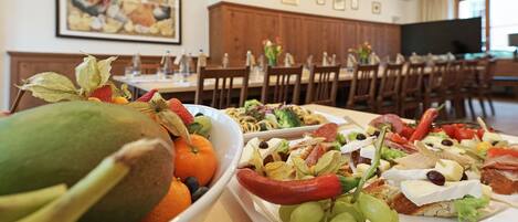 A dining area with a variety of food platters, including fruits, vegetables, and cheeses. There are wooden chairs and tables, and a television is visible in the background.