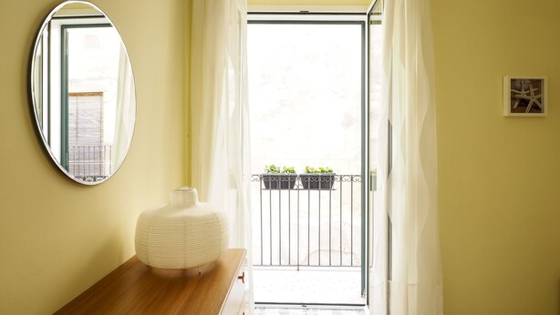 Une pièce avec une table en bois, un vase blanc, un miroir rond et une vue sur un balcon agrémenté de plantes en pot.
