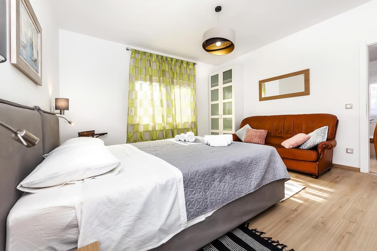 Quarto de hotel bem organizado, com cama, sofá, abajur e janela com cortinas.
