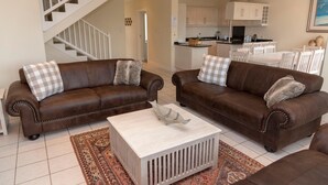 A living room with two brown leather sofas, a white coffee table, and a patterned rug. A staircase is visible in the background leading to a kitchen area. - Villa 17 (Knysna)