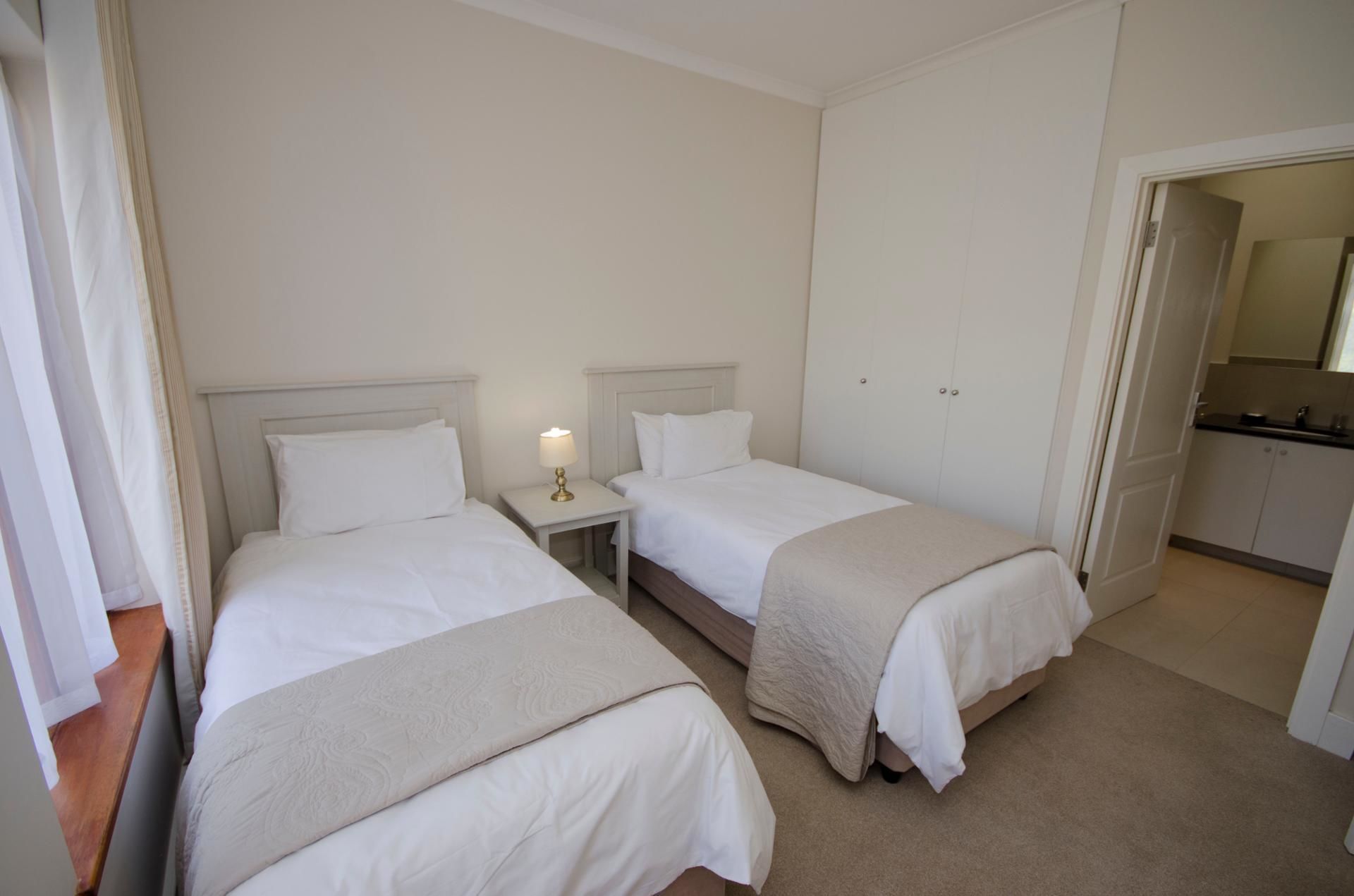 A hotel room with two single beds, a nightstand with a lamp, a wardrobe, and a small kitchenette.