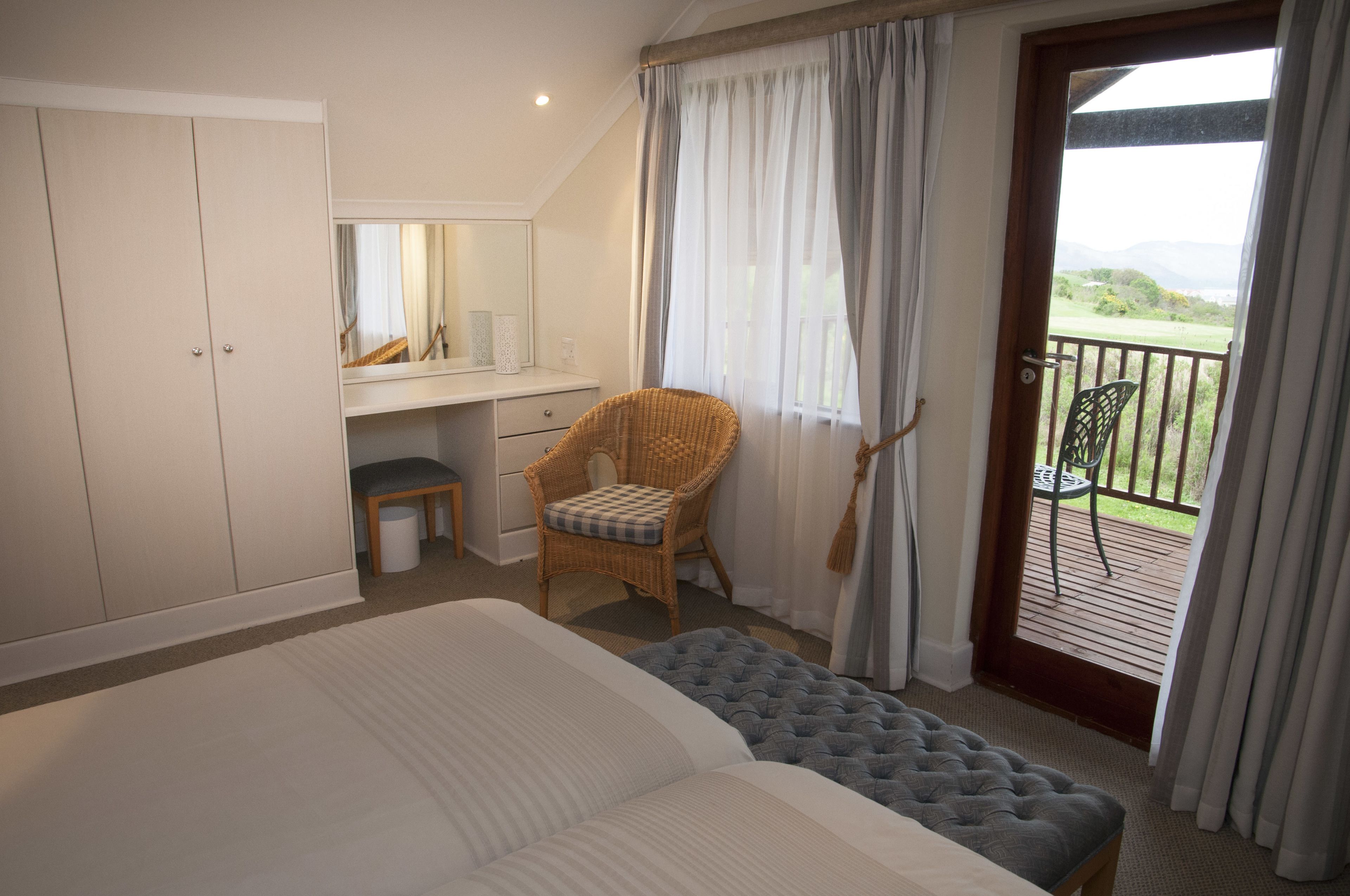 A bedroom with a bed, a chair, a desk, a mirror, and a balcony with a view.