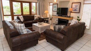 A living room with two brown leather sofas, a white coffee table, a flat-screen TV, and a fireplace. - Villa 17 (Knysna)