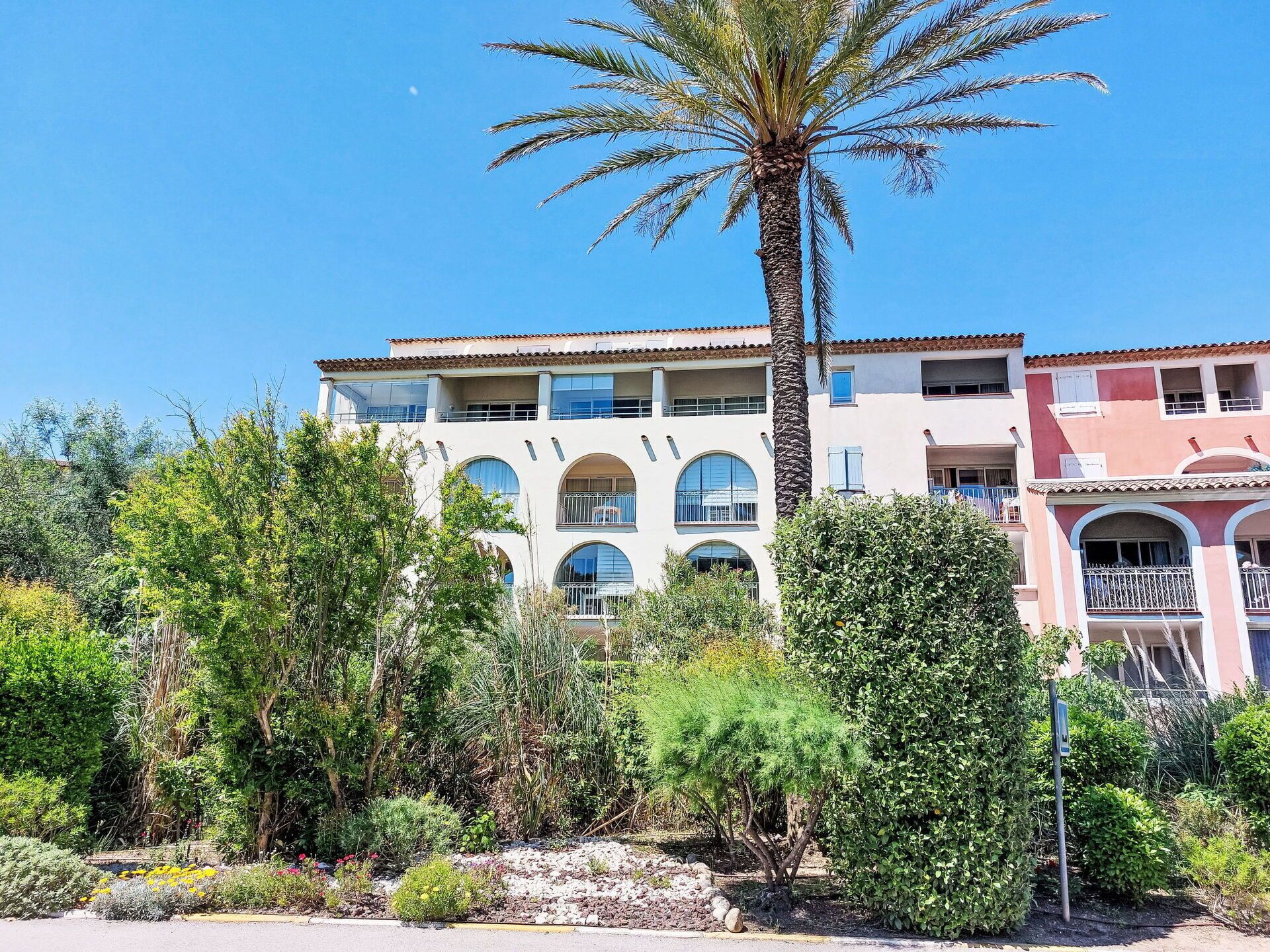 Un edificio de varios pisos con balcones, rodeado de exuberante vegetación y una palmera.