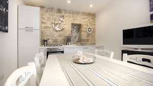A modern kitchen with a stone wall, white cabinetry, and a dining area with a long table and white chairs. - Just 100 meters from one of the most beautiful and renowned beaches of the Ragusa’s coast (Marina di Ragusa)