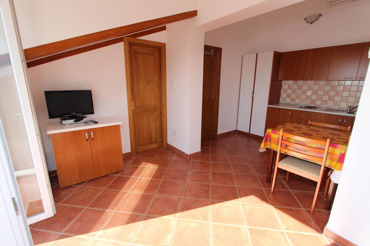 A compact kitchen with a tiled floor, wooden cabinets, a television, and a dining table with a colorful tablecloth.