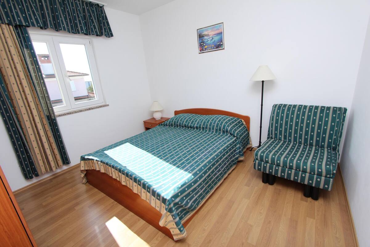 A bedroom with a bed, a chair, a nightstand, a lamp, and a window with curtains.