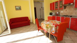 A compact kitchen with red cabinets, a dining table with a floral tablecloth, and a red sofa. - Apartments Elida (Rovinj)