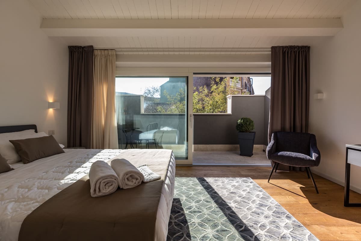 A modern hotel room with a large bed, a small desk, a chair, and a balcony with a view of the city.
