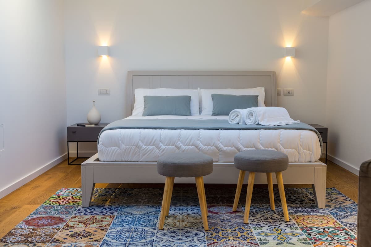A neatly made bed with a white duvet and two pillows, a bedside table with a vase, and a colorful patterned rug.