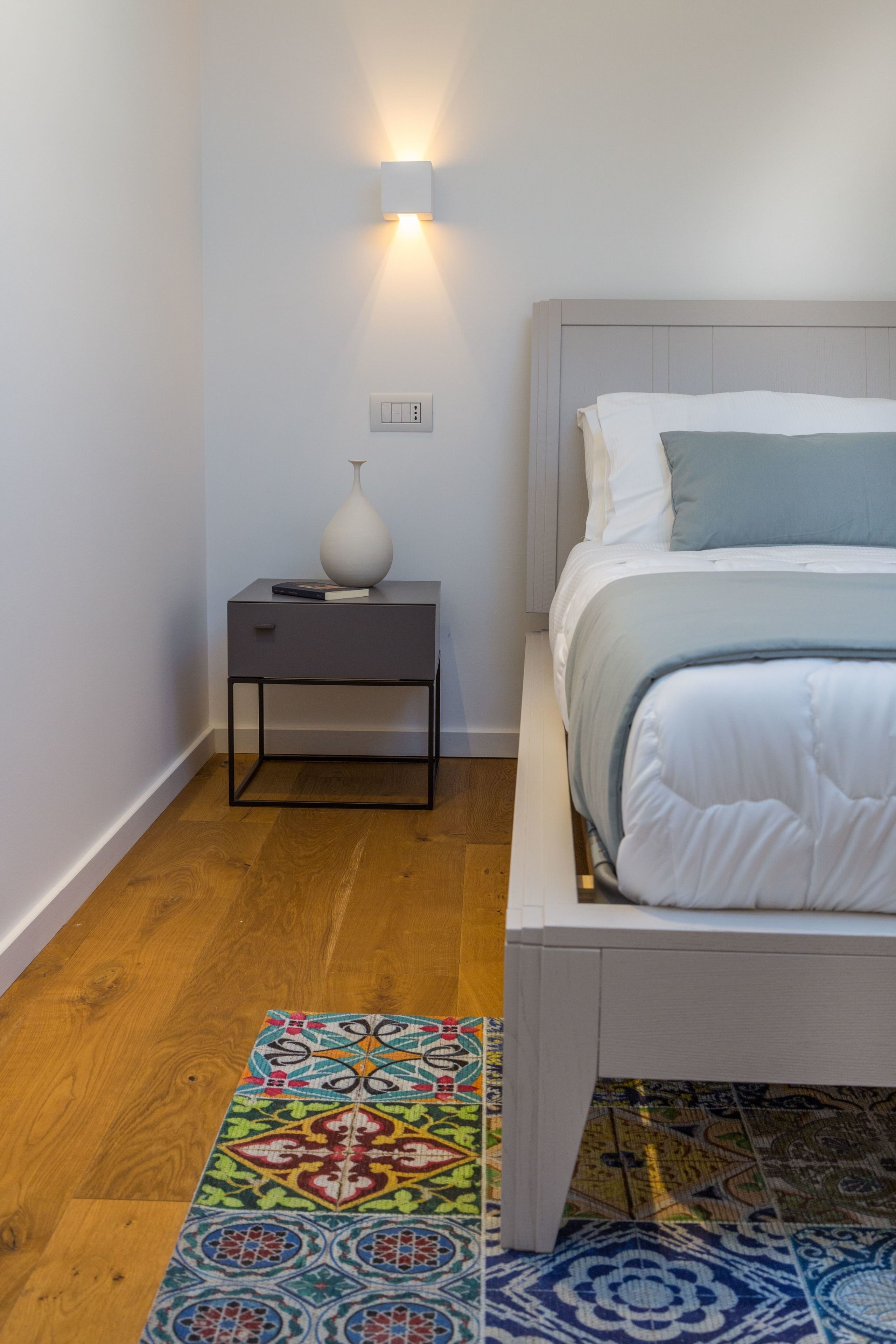 A bedroom with a bed, a bedside table with a lamp, a nightstand, and a colorful rug.