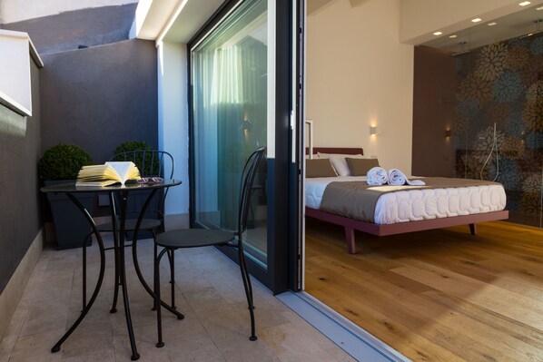 A modern bedroom with a bed, a small table with chairs, and a glass door leading to a balcony. - Plebis - Camere & Suites in Sicilia nel centro storico di Catania (Catania)