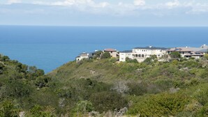 A coastal property with multiple buildings, including a large house with a sloped roof and solar panels, situated on a hillside with dense greenery and a view of the ocean. - Villa 3 (Knysna)