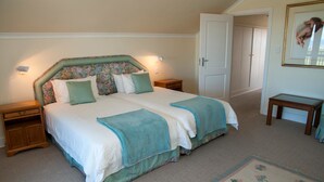 A bedroom with a double bed, floral headboard, bedside tables, a nightstand, a small wooden table, a framed picture, and a carpeted floor. - Villa 3 (Knysna)
