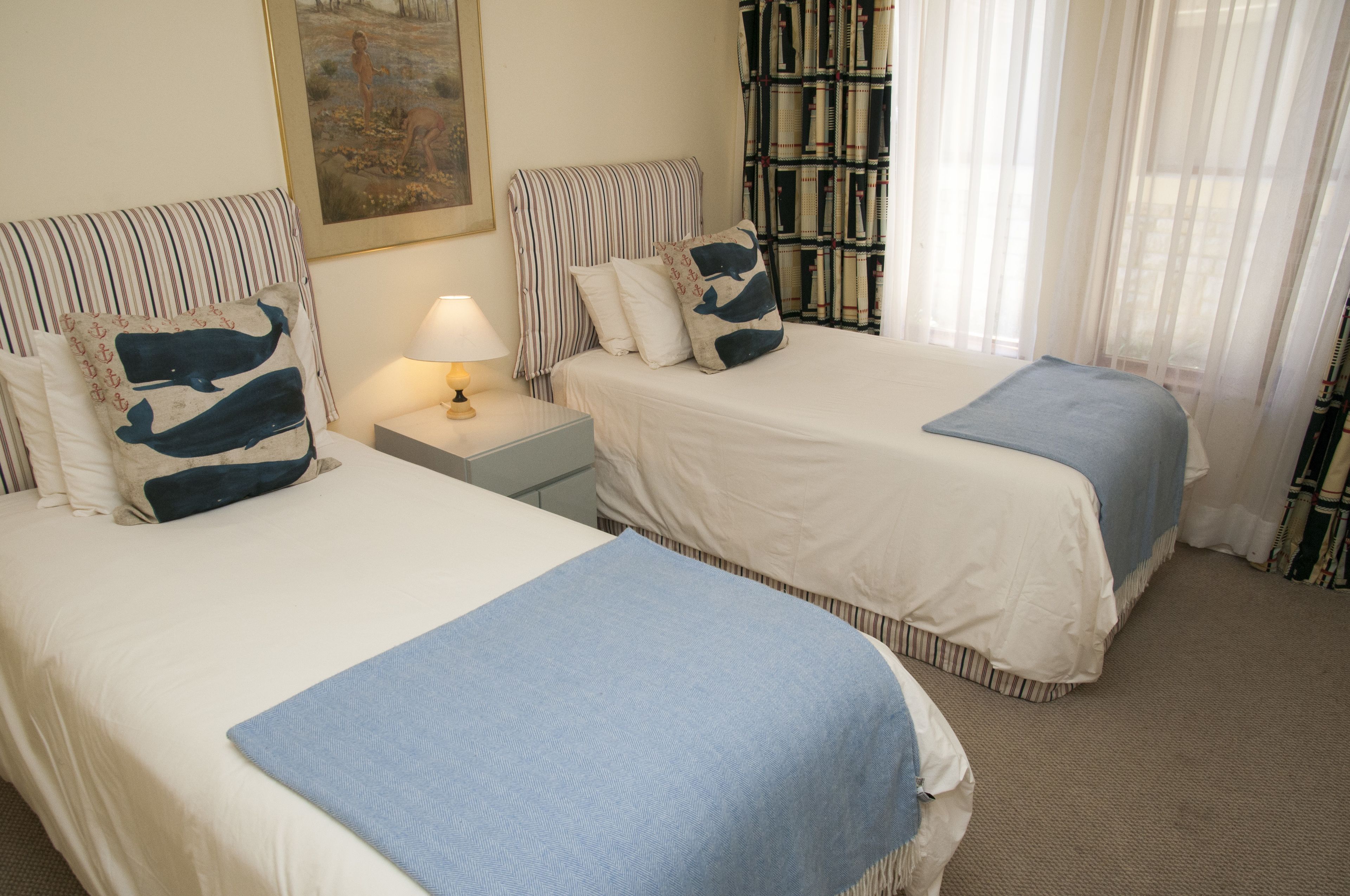 A hotel room with two beds, a nightstand with a lamp, a painting on the wall, and sheer curtains.
