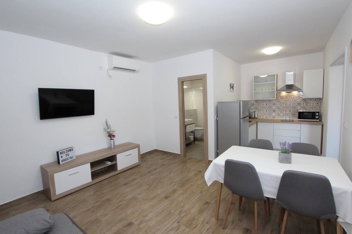 A modern living room with a flat-screen TV, a wooden entertainment unit, and a dining area with a table and chairs.