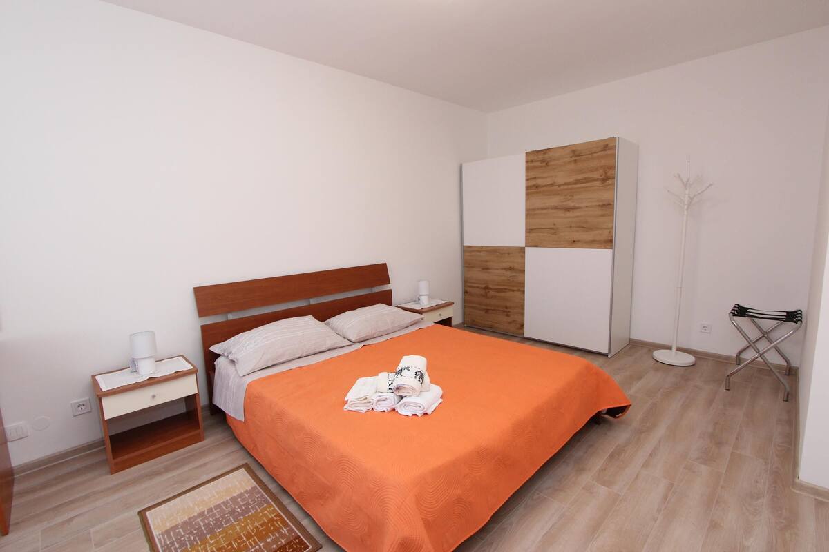 A neatly arranged bedroom with a wooden headboard, a single bed with an orange bedspread, a bedside table with a lamp, and a wardrobe with a wooden panel design.