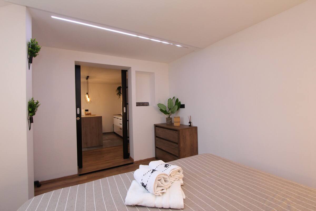 A modern bedroom with a bed, a wooden dresser, a plant, and a bathroom visible through an open door.