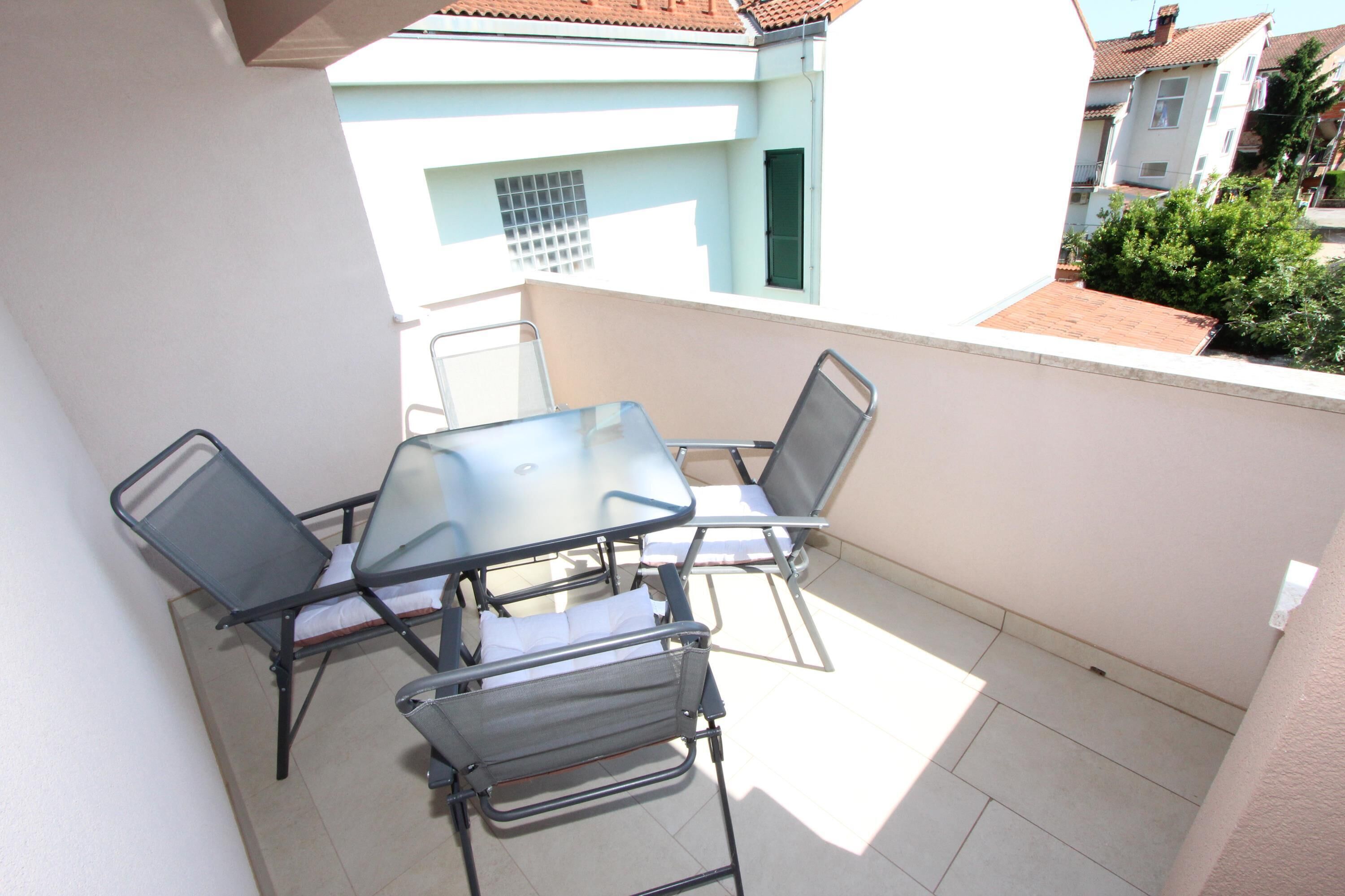A balcony with a glass table and metal chairs, a tiled floor, and a view of neighboring buildings.
