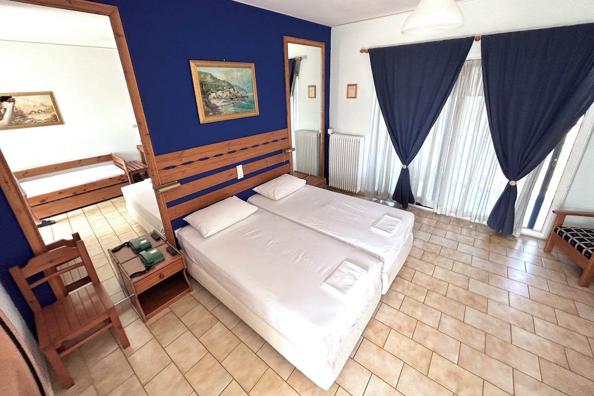 A double bed with white linens, a wooden headboard, a bedside table with a lamp, a chair, and a painting on the wall.