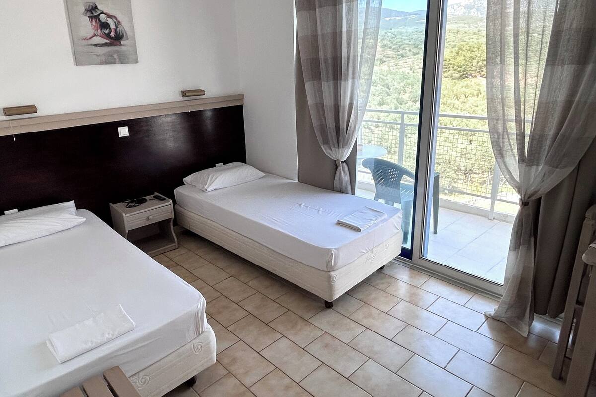 A hotel room with two beds, a wooden headboard, a small table with a lamp, and a balcony with a view of the countryside.
