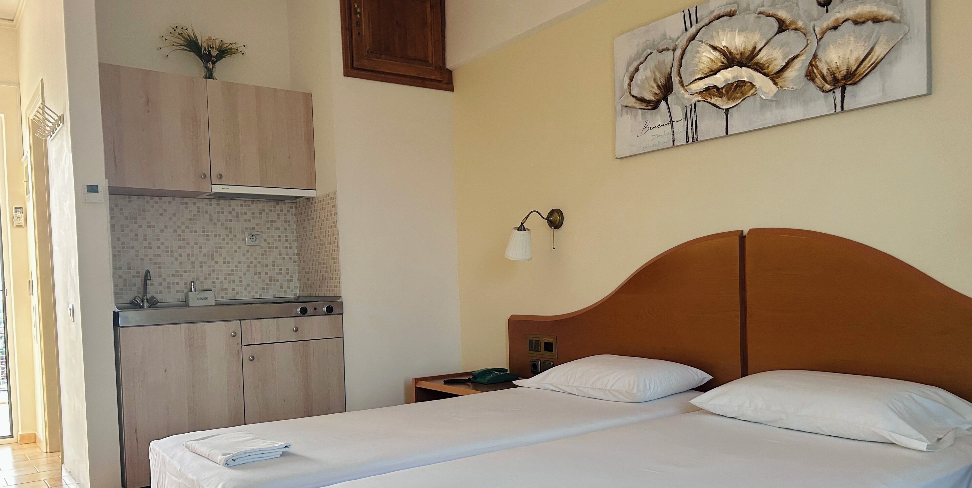 Une chambre avec une tête de lit en bois, un lit recouvert de linge de maison blanc, une table de chevet avec une lampe, et un coin cuisine doté de meubles en bois et d’un dosseret carrelé.