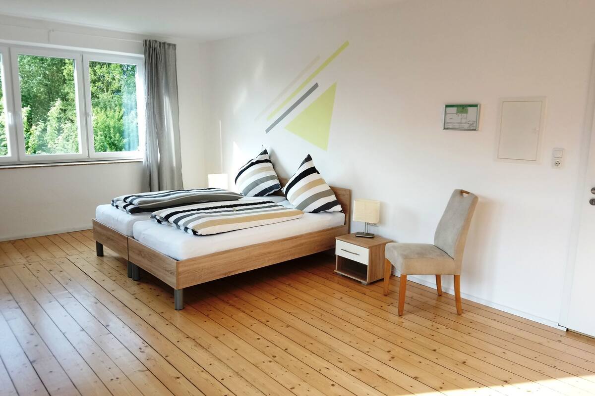 A bedroom with a wooden bed, a nightstand, a chair, a window with curtains, and a wall-mounted light.