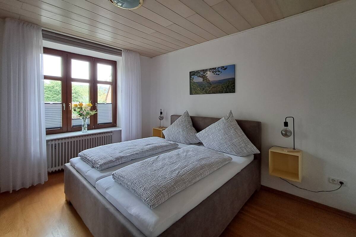 A bedroom with a large bed, a window with curtains, a radiator, a bedside table with a lamp, and a wall-mounted picture.