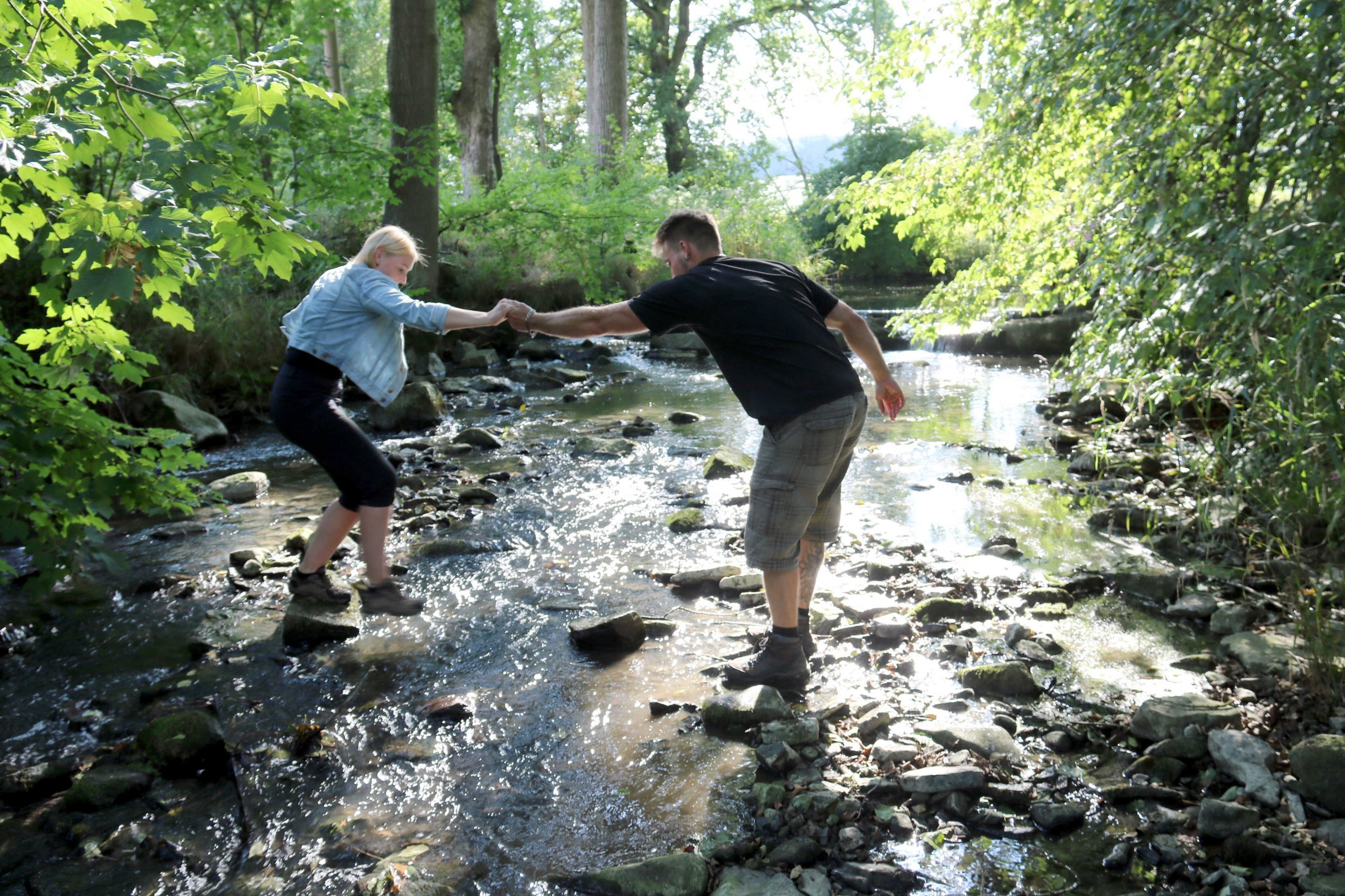 Dos personas cruzando un arroyo pedregoso en un área boscosa.